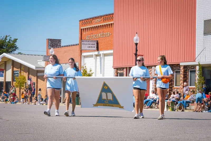 Young women carry a white banner bearing the Biblesta emblem, a gold triangle around an open Bible.