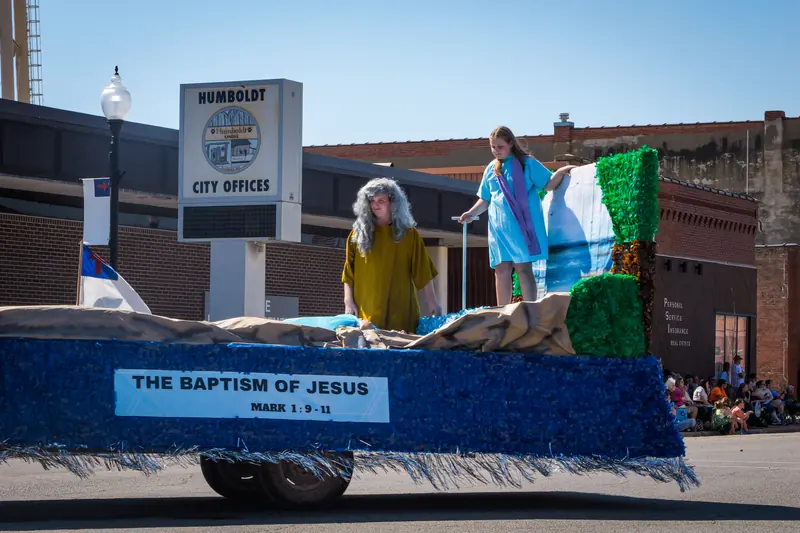 Costumed figures stand at the river's edge on a float titled The Baptism of Jesus.