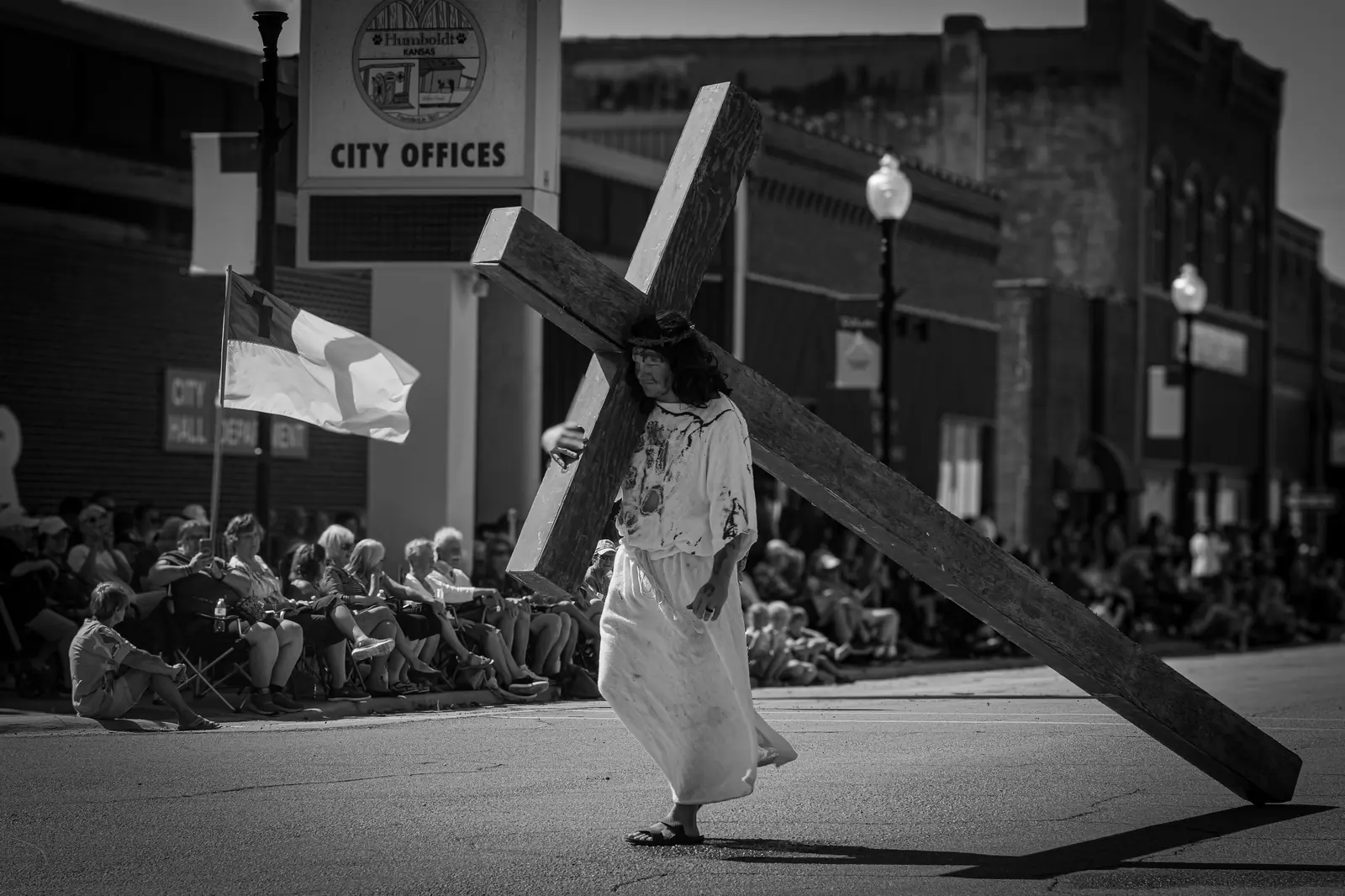 In black and white, a man portraying Jesus drags a wooden cross past rows of seated spectators downtown.